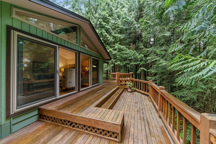A green home has a spacious wooden deck that looks out to a wooded area.